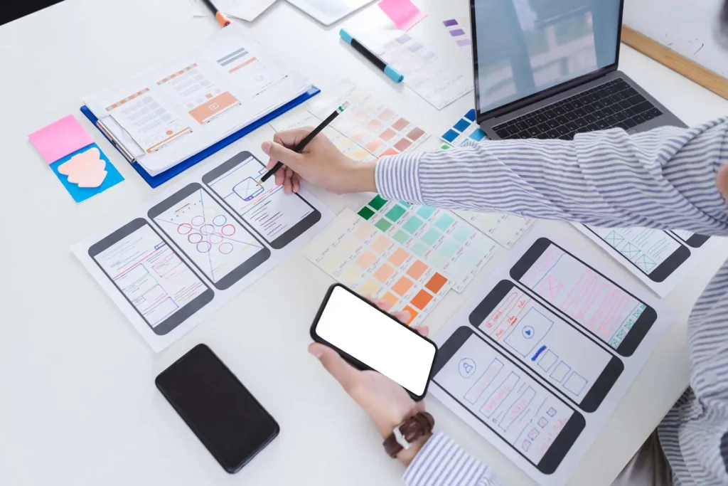 นักออกแบบกำลังสเก็ตช์ Wireframe หน้าจอมือถือ พร้อมแล็ปท็อป ชุดสี และเอกสารดีไซน์ สื่อถึงการออกแบบ UX UI การพัฒนาเว็บไซต์ Responsive และประสบการณ์ผู้ใช้
