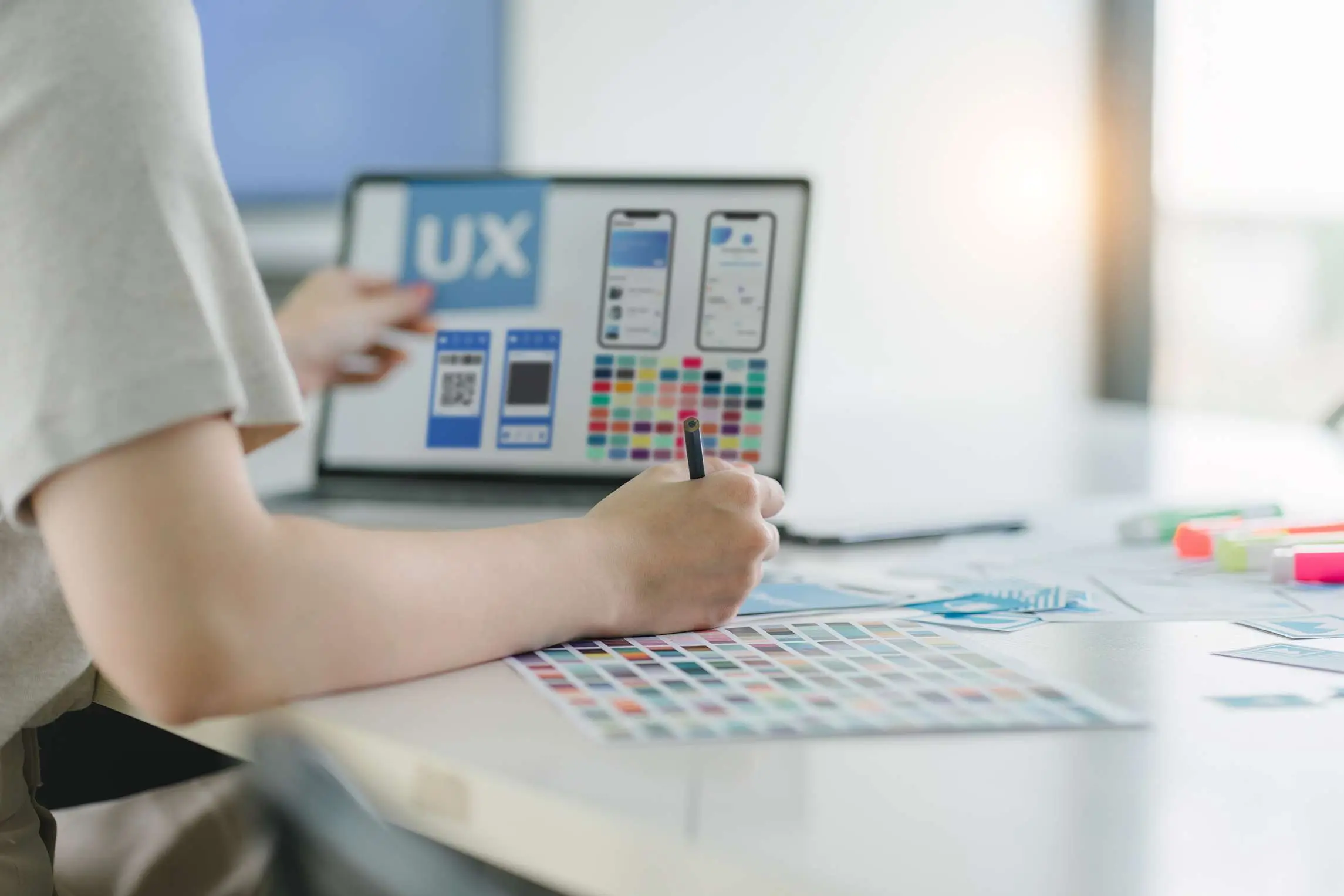 ภาพนักออกแบบกำลังวางแผน UX UI พร้อม Wireframe หน้าจอแอป และชุดสีงานดีไซน์ สื่อถึงการออกแบบเว็บไซต์ แอปพลิเคชัน และประสบการณ์ผู้ใช้